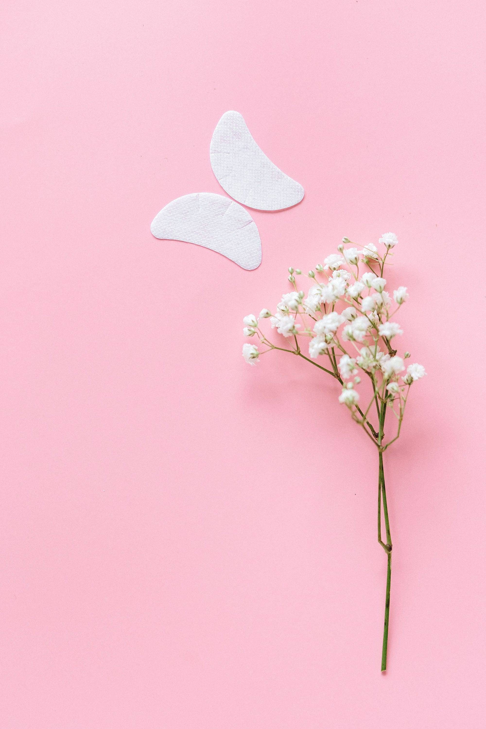 Hydrating under-eye patches next to a sprig of baby’s breath on a pink background