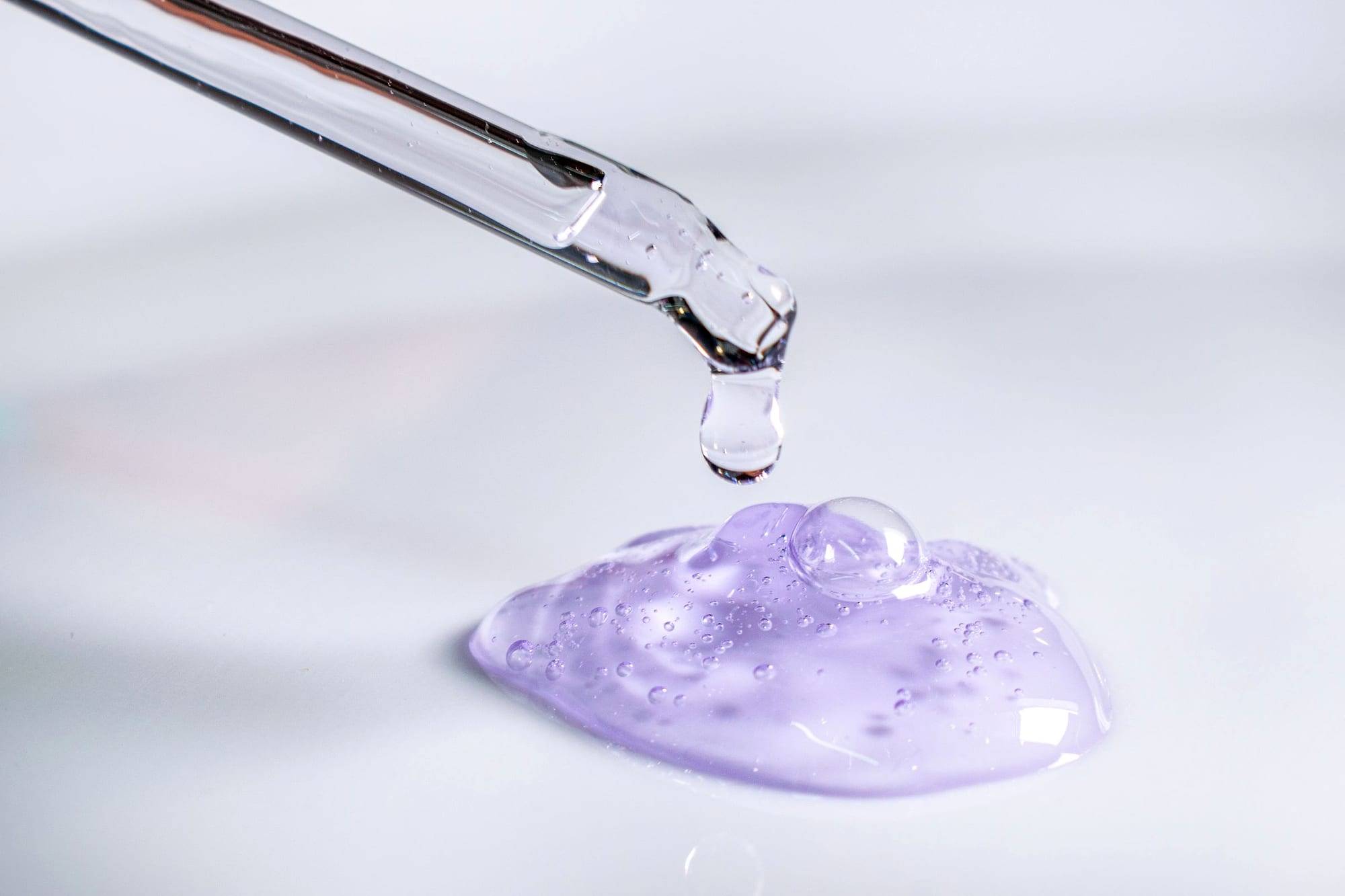 Pipeta dejando caer una gota de gel cosmético lila sobre una superficie blanca