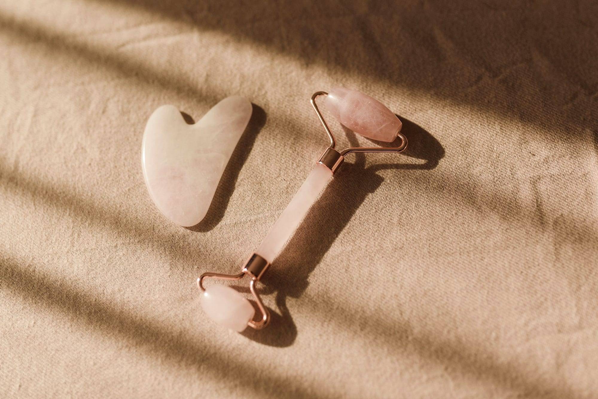 Rodillo facial y piedra gua sha de cuarzo rosa sobre una tela beige con luz suave